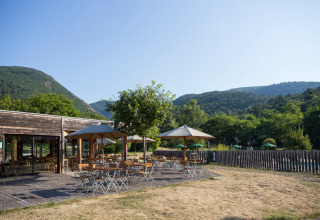 Udendørs caféområde med parasoller, borde og stole i Village Huttopia Dieulefit, Auvergne-Rhône-Alpes, omgivet af bjerge.