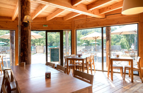 Interior de restaurante de madera en Village Huttopia Dieulefit con vistas a terraza exterior y sombrillas.