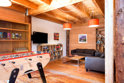 Sala luminosa con paredes de madera, futbolín, estanterías con libros y sofás en Huttopia Dieulefit.