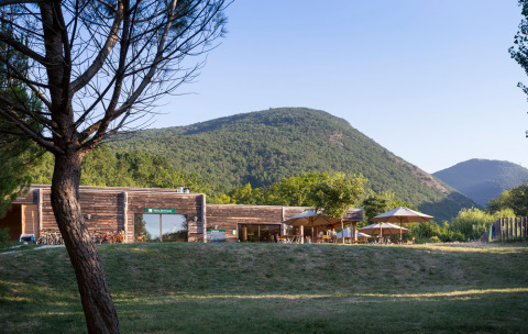 Modern wooden cabin with sun umbrellas in green mountain scenery at Village Huttopia Dieulefit, France.