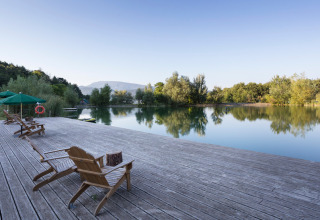 Peaceful lakeside view with wooden deck, lounge chairs, and green umbrellas at Village Huttopia Dieulefit holiday park.
