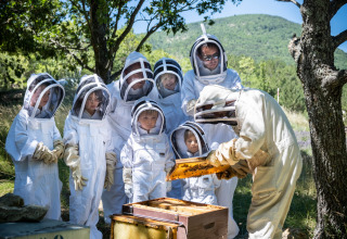 Children and adults in beekeeping suits watch a beekeeper examine a hive at Huttopia Dieulefit, France.