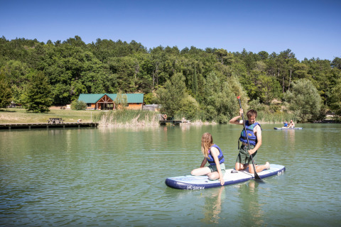 Personas practicando paddle surf en un lago tranquilo en Village Huttopia Dieulefit, rodeados de naturaleza y cabañas.