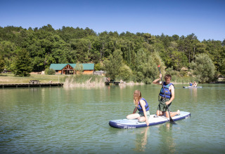 Persone che fanno paddleboard su un lago calmo a Village Huttopia Dieulefit, circondati da foresta e chalet.