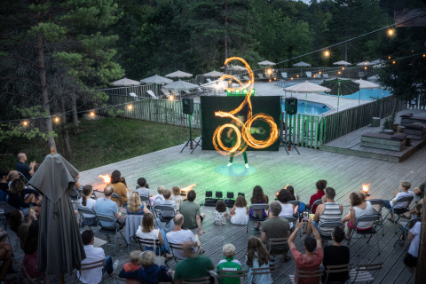 Publiek kijkt naar een vuurshow op een houten terras met zwembad bij Village Huttopia Lanmary, Frankrijk.