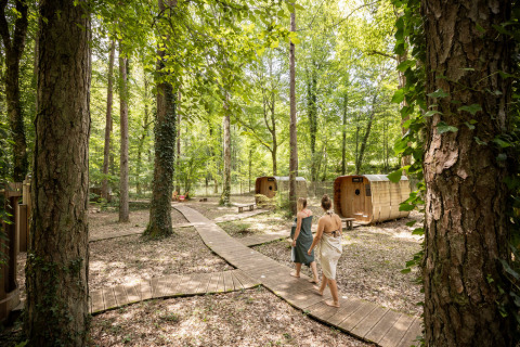 Dos personas caminan por un sendero de madera en el bosque junto a cabañas en Village Huttopia Lanmary, Francia.