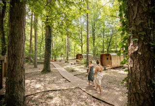 Two people walk on a wooden path through the forest near small cabins at Village Huttopia Lanmary, France.