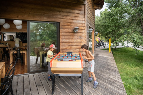 Dos niños juegan futbolín en una terraza de madera junto a una cabaña en Huttopia Lanmary, Francia.