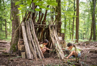 Zwei Kinder spielen vor einer Holzhütte im grünen Wald des Village Huttopia Lanmary in Frankreich.