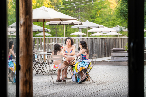 Drie personen in zwemkledij aan een tafel op het terras van Village Huttopia Lanmary, Frankrijk, onder parasols.