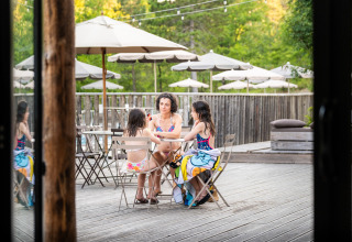 Three people in swimsuits sit at an outdoor table with umbrellas at Village Huttopia Lanmary, France holiday park.