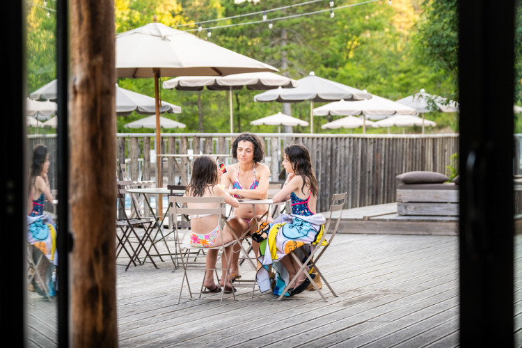 Trois personnes en maillot sur une terrasse avec parasols à Village Huttopia Lanmary, parc de vacances en France.