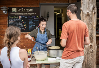 En kvinde laver pandekager foran caféen på Village Huttopia Lanmary med gæster, der venter på servering.