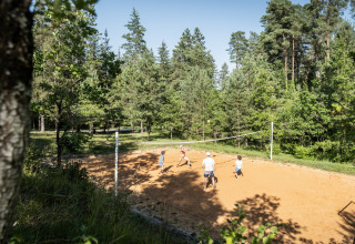 Folk spiller volleyball på en udendørs bane omgivet af træer i ferieparken Huttopia Lanmary, Frankrig.