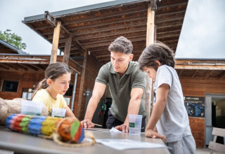 Ein Mann und zwei Kinder arbeiten gemeinsam an einem Tisch im Village Huttopia Lanmary, Nouvelle-Aquitaine, Frankreich.