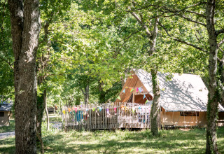 Safari tent in a lush forest, colorful bunting hanging on the fenced deck, sunlight streaming through trees.