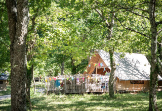 Safari tent in a lush forest, colorful bunting hanging on the fenced deck, sunlight streaming through trees.