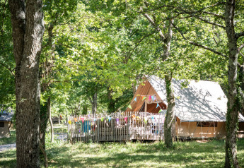Safari tent in a lush forest, colorful bunting hanging on the fenced deck, sunlight streaming through trees.