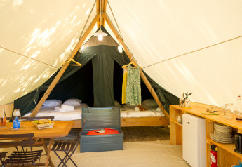 Interior view of a safari tent featuring a dining area, kitchenette, two beds, and sunlit ambiance.