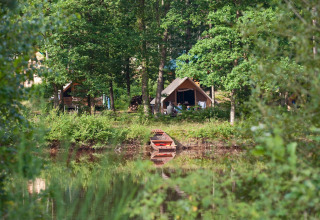 Safari-telt ved en sø omgivet af frodig skov, en lille robåd ligger forankret ved bredden om sommeren.