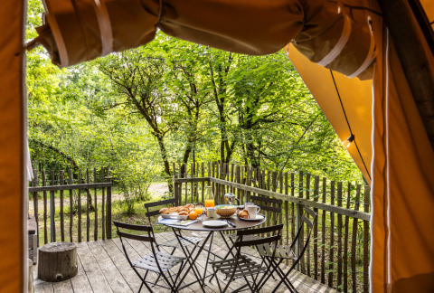 Uitzicht vanuit een safaritent op een houten terras met ontbijt en vier stoelen in een groen bos.