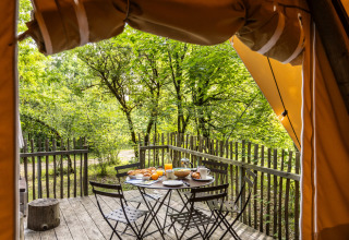 Uitzicht vanuit een safaritent op een houten terras met ontbijt en vier stoelen in een groen bos.