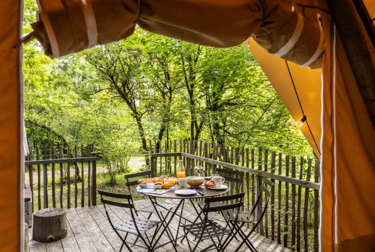 View from a safari tent opening to a wooden deck with breakfast and four chairs in a lush forest.