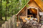 Safari-telt i skoven med overdækket træterrasse, udendørs spiseplads og campingkøkken i naturen.