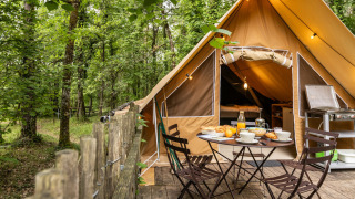 Tenda safari nel bosco con terrazza in legno, tavolo apparecchiato e cucina da campeggio esterna.