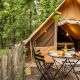 Safari-telt i skoven med overdækket træterrasse, udendørs spiseplads og campingkøkken i naturen.