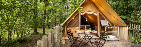 Safari tent in the woods, with wooden deck, outdoor breakfast table, and camping kitchen in nature.
