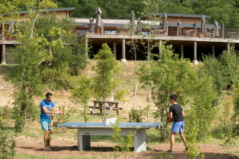 Dos personas juegan al tenis de mesa al aire libre rodeados de árboles, con cabañas de madera detrás en Francia.