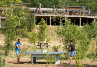 Zwei Menschen spielen im Freien Tischtennis vor Bäumen, mit Holzgebäuden im Hintergrund in Frankreich.
