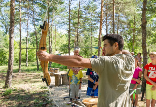 Ein Mann bringt Kindern im Ferienpark Village Huttopia Sud-Ardèche im Wald das Bogenschießen bei.