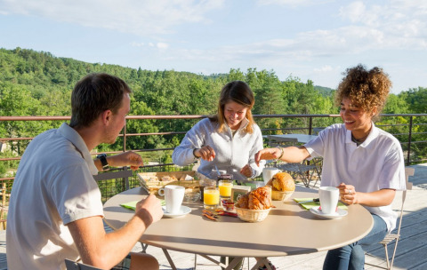Tres personas desayunan al aire libre en una terraza con vistas verdes en Village Huttopia Sud-Ardèche, Francia.