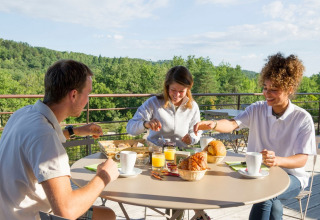 Tres personas desayunan al aire libre en una terraza con vistas verdes en Village Huttopia Sud-Ardèche, Francia.