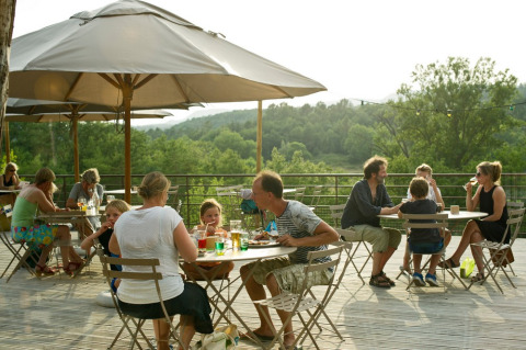 People dine and relax on a sunny outdoor terrace overlooking the forest at Village Huttopia Sud-Ardèche.