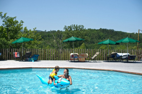 Une mère et un enfant jouent avec un dauphin gonflable bleu dans la piscine extérieure d’Huttopia Sud-Ardèche.