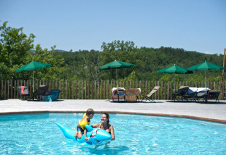 En mor og et barn leger i en udendørs pool med en blå oppustelig delfin i ferielandsbyen Huttopia Sud-Ardèche.