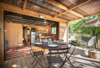 Essbereich auf einer Holzterrasse mit offener Küche in einer Hütte im Village Huttopia Sud-Ardèche, Frankreich.