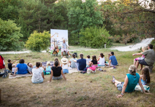 En gruppe familier sidder udendørs på tæpper og kigger på en fortæller ved Village Huttopia Sud-Ardèche.
