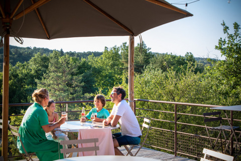 En familie nyder en afslappet frokost på en terrasse med udsigt til grønne træer i Village Huttopia Sud-Ardèche.