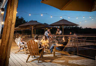 Udendørs terrasse ved skumring på Village Huttopia Sud-Ardèche, hvor gæster slapper af under træparasoller.