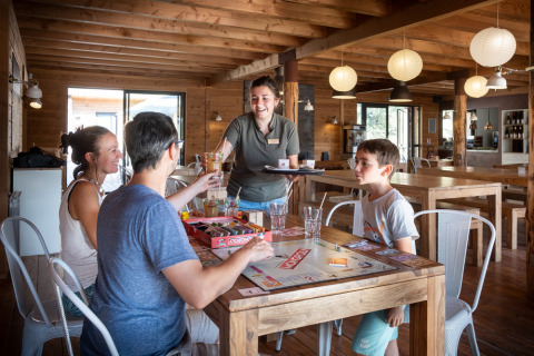 Familia juega a un juego de mesa y recibe bebidas en Village Huttopia Sud-Ardèche, Auvergne-Rhône-Alpes.