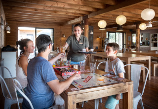 Famille jouant à un jeu de société et recevant des boissons au Village Huttopia Sud-Ardèche, Auvergne-Rhône-Alpes.