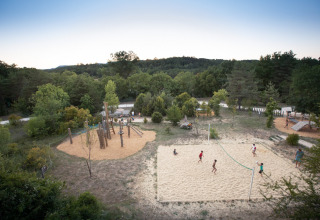 Børn leger volleyball på en sandbane og på legeplads i Village Huttopia Sud-Ardèche, omgivet af skov.