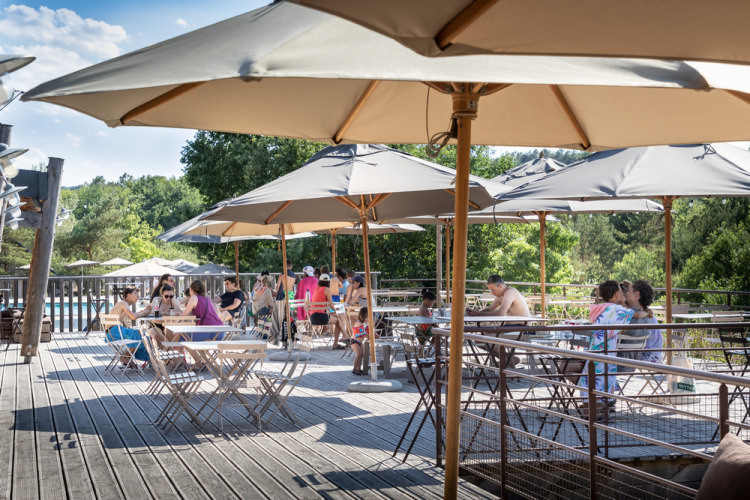 Terrasse extérieure avec parasols et visiteurs au Village Huttopia Sud-Ardèche en Auvergne-Rhône-Alpes, France.