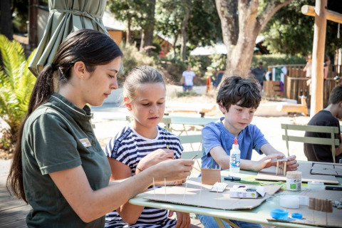 Children and an adult do outdoor crafts with glue and cardboard at Huttopia La Forêt de Janas, France.