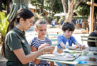 Kinderen en een begeleidster knutselen buiten met lijm en karton bij Huttopia La Forêt de Janas, Frankrijk.