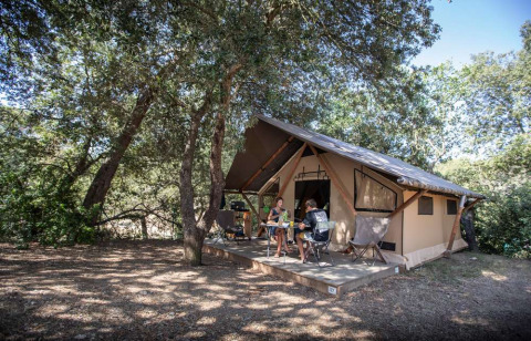 En familie nyder en ferie foran et glampingtelt under træerne på Huttopia La Forêt de Janas i Frankrig.
